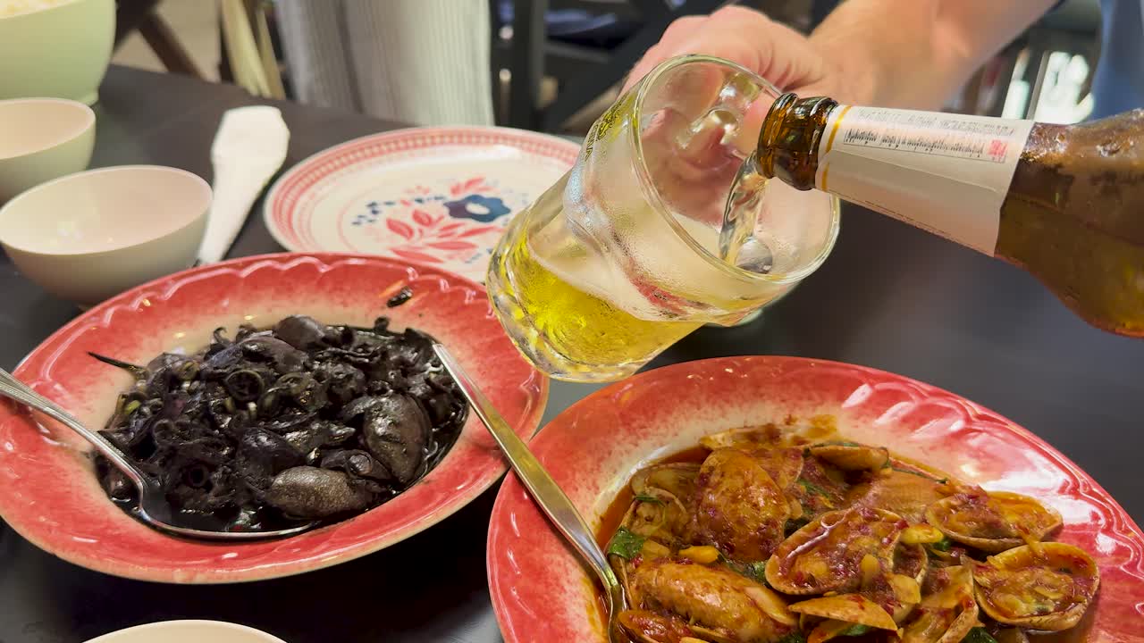 Hand pours light beer into a glass with ice next to plates of spicy clams and black mussels in a brightly lit restaurant setting