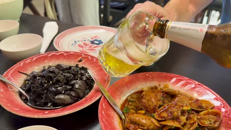 hand pours light beer into a glass with ice next to plates of spicy clams and black mussels in a brightly lit restaurant setting