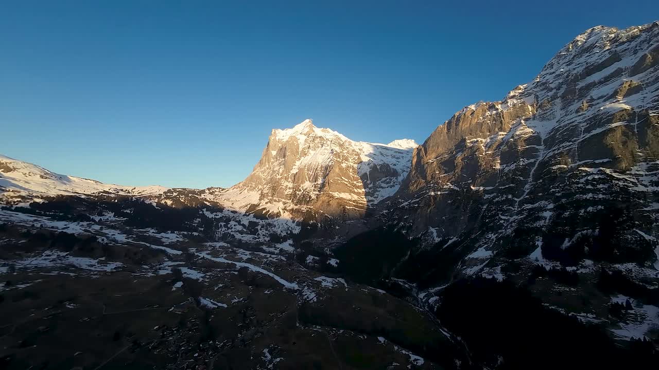 Panoramic aerial view of mountain peaks in Alps. Glacial gorge and steep, rugged cliff faces with snow covered ridge and golden sunlight. Pan right.