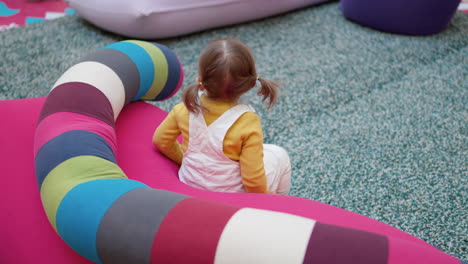 a blond hair 3-year-old girl toddler in ponytails sitting on a soft pink couch with a colorful long pillow at the play area inside a shopping mall