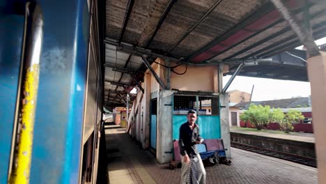 a man dashes along the platform at kandy station in sri lanka, trying to catch a departing train. a woman sits nearby on a bench, observing the scene. the setting is lively and dynamic.