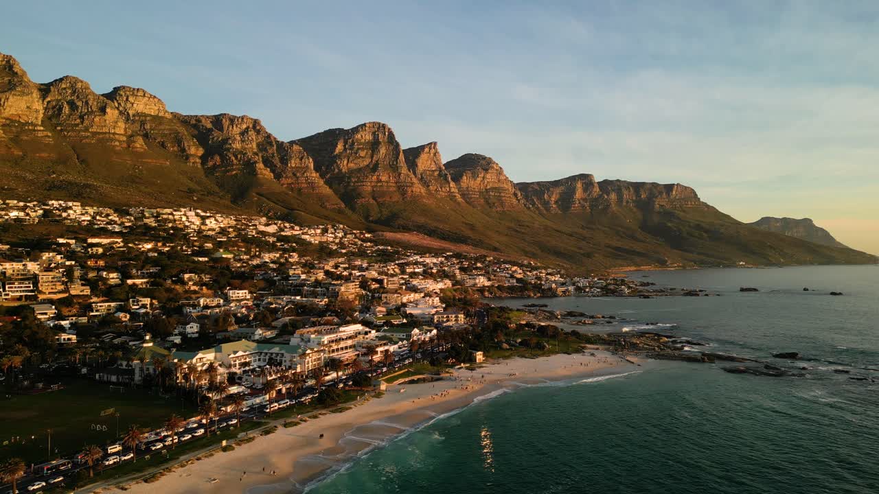 Letecké záběry dronem odlétající od Camps Bay v Kapském Městě v Jihoafrické republice během zlaté hodiny, ukazující pobřežní krajinu a teplé světlo západu slunce.