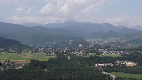 Aerial-panorama-view-of-Bled-Village-with-green-Valley-fields,-forest-and-mountain-range-during-mountain-chain-in-Background