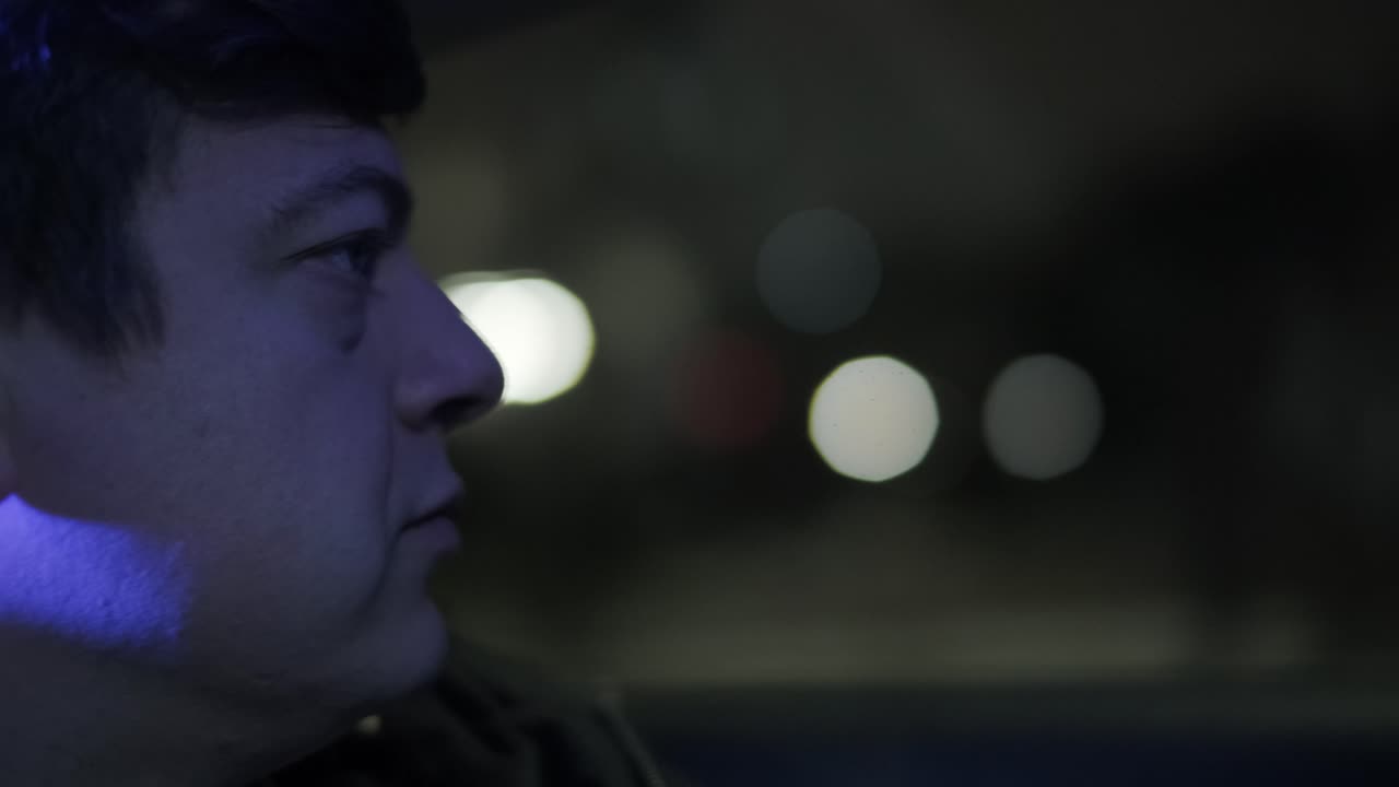 멋진 젊은 사업가의 느린 동작 클로즈업은 밤에 도시의 중심에서 운전자와 함께 현대적인 차로 여행하는 동안 밖을 바라보고 있습니다.