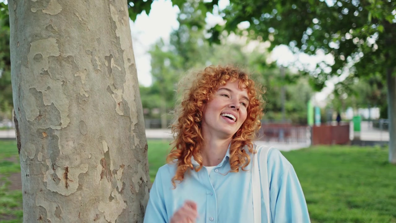Giovane donna felice con capelli rossi ricci appoggiata a un albero in un parco.