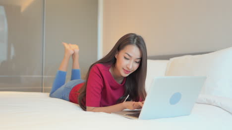 woman lying on the bed with her laptop in front of her and typing message on the keyboard, happy smiling expression on her face, slow-motion perspective view