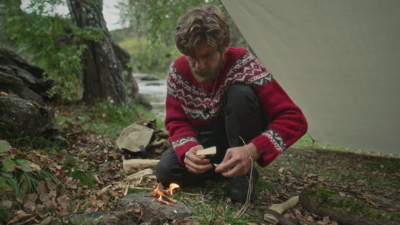 Survivalist Tending Small Campfire under Tarp Shelter in Forest
