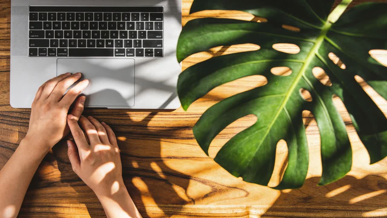 Skiftende sollys, hender legger seg på sølvfarget laptop på trebord, Monstera-bladskygger for arbeid.