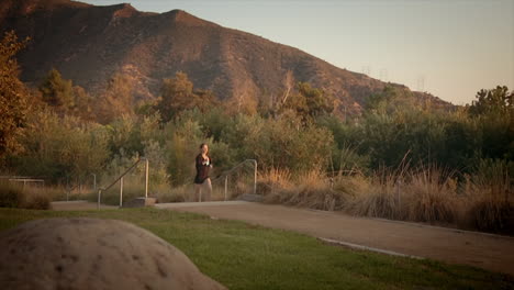 Wide-angle-dolly-shot-of-a-beautiful-slim-caucasian-woman-jogging-along-a-path-amid-the-sun-setting-against-the-mountain-in-the-background