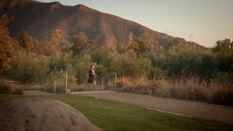 wide angle dolly shot of a beautiful slim caucasian woman jogging along a path amid the sun setting against the mountain in the background