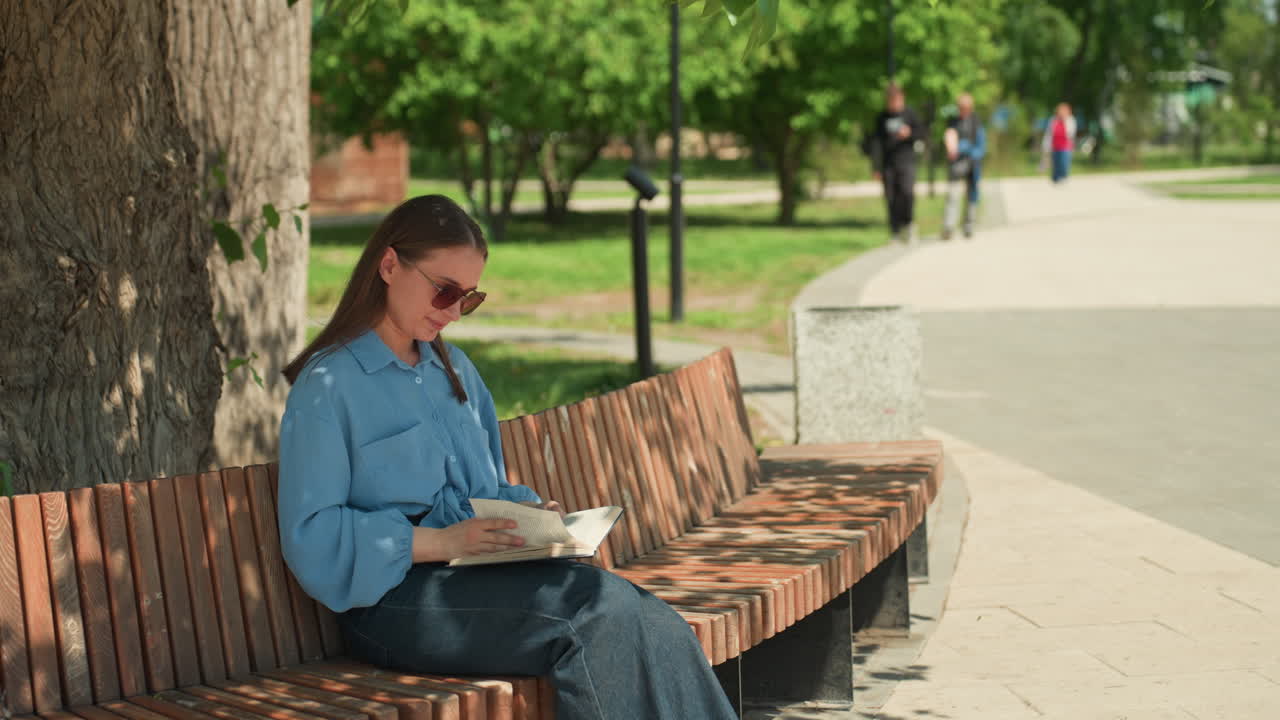 선글라스를 끼고 나무 그늘 아래에서 책을 읽는 편안한 표정의 백인 여성, 빛이 얼룩덜룩한 여름 공원 풍경, 캐주얼한 데님과 산들바람에 흩날리는 자세, 조용한 여가와 책장을 넘기며 마음을 달래는 모습