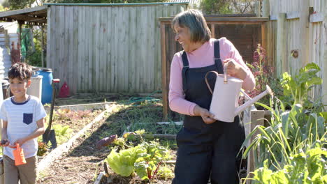 Feliz-Abuela-Birracial-Y-Nieto-Regando-Plantas-En-Un-Jardín-Soleado,-Cámara-Lenta