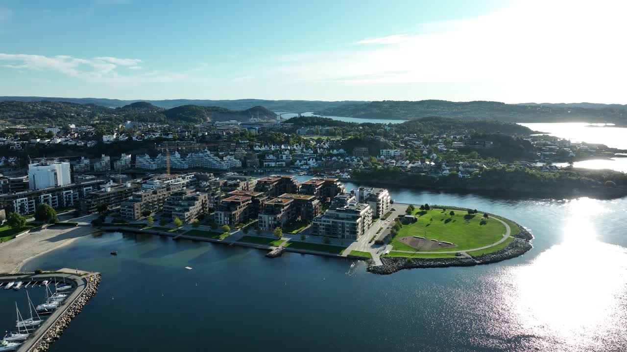 Bright sunny day showcases Kristiansand in Norway with modern architecture, a serene marina, and lush green areas along the waterfront. Scenic hills rise in the background