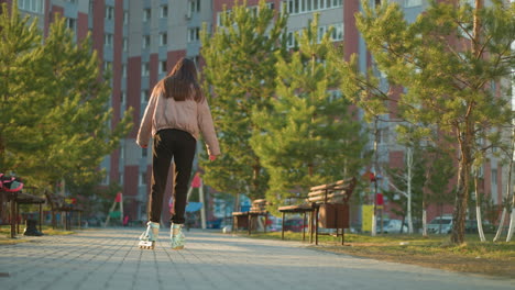 a girl in a peach jacket and black trousers is captured from the back as she rollerblades through a park. rows of trees and benches, with tall residential buildings in the background