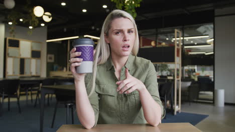 caucasian businesswoman having a video chat drinking a cup of coffee in modern office