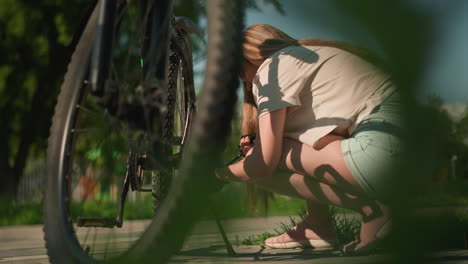 close-up of lady squatting beside her bicycle, intently pumping air into tire using green nozzle on a sunny day, bicycle is in focus, while background with fence and trees softly blurred