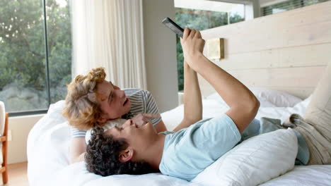 Feliz-Pareja-Gay-Diversa-Acostada-En-La-Cama-Y-Usando-Un-Teléfono-Inteligente-En-Casa,-Cámara-Lenta