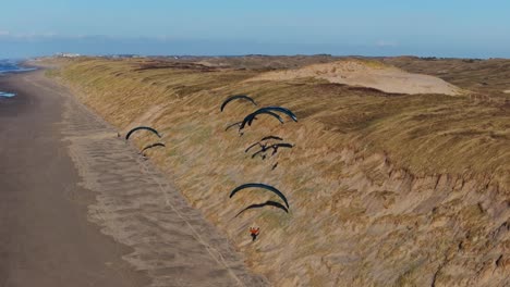 Grupo-De-Parapentes-Se-Elevan-A-Lo-Largo-De-Las-Laderas-De-Dunas-De-Arena-Cubiertas-De-Hierba-De-La-Playa-De-Castricum,-Países-Bajos