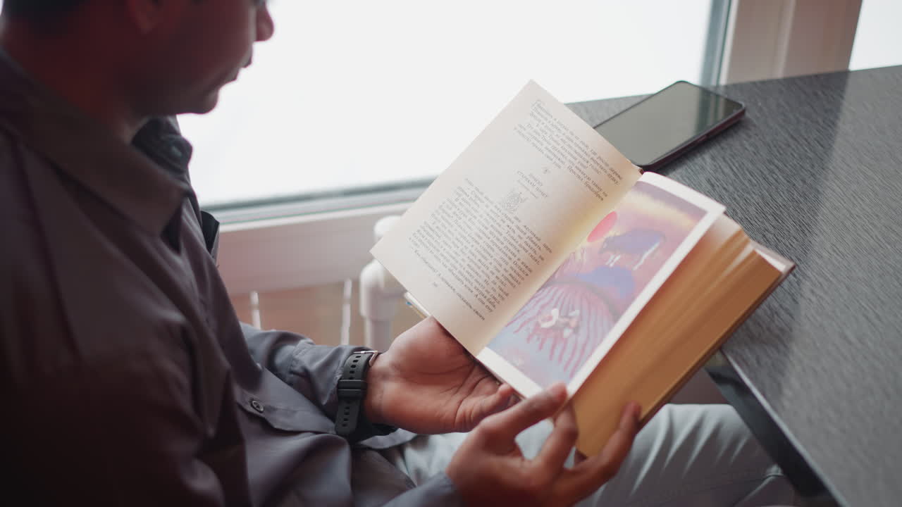 Student sitting near window in red booth on snowy day reading open book with colorful illustration, holding pages with both hands, smartphone on table, relaxed posture, indoor learning environment