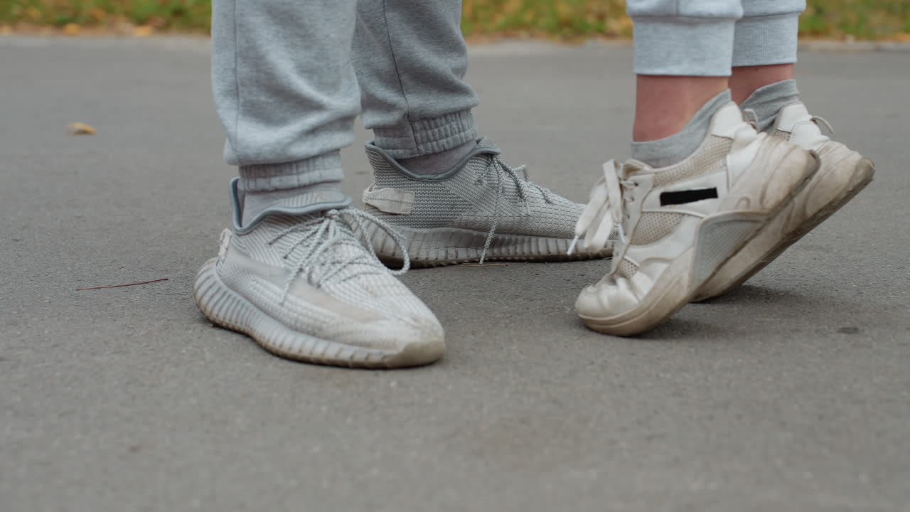 Side leg view of two people wearing joggers and sneakers standing on pavement as one person raises feet slightly in playful motion with blur background