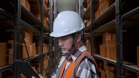 close up side view of asian male engineer with safety helmet working on a tablet while standing in the warehouse with shelves full of delivery goods