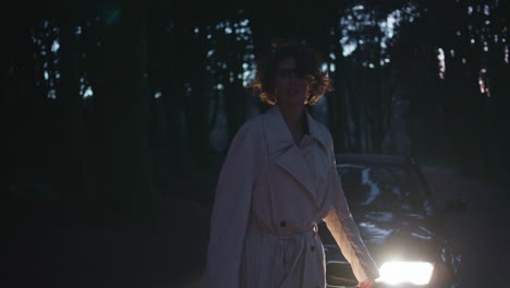 woman standing car headlights in dark forest backdrop. elegant model at night