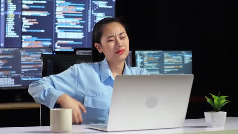 asian female programmer having a backache while writing code by a laptop using multiple monitors showing database on terminal window desktops in the office