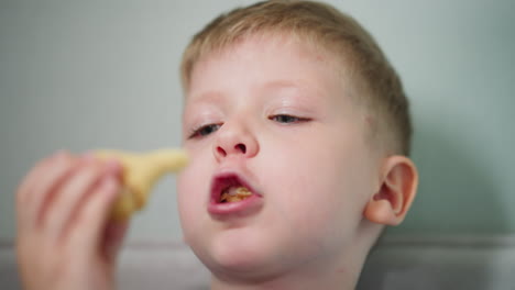 close up of little boy with blond hair wearing blue shirt looking up thoughtfully while eating snack indoors capturing peaceful moment of childhood innocence with soft lighting and calm background