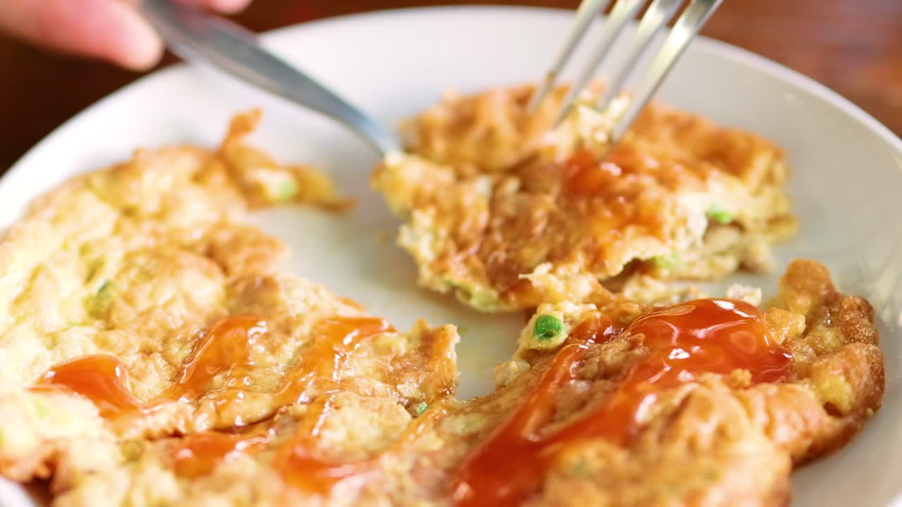 A fork and spoon cut into a ketchup-covered omelet on a white plate.