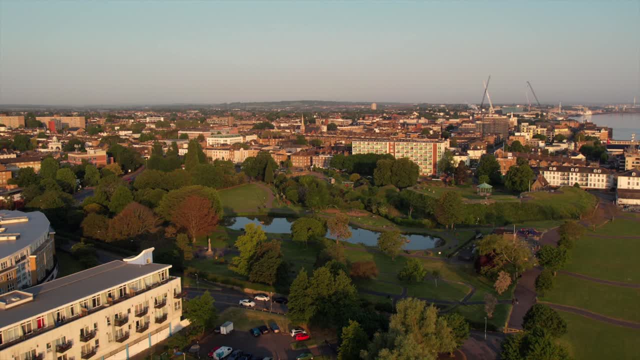 Gravesend cityscape at sunrise, featuring Gordon Lake and a public park. The scene includes residential buildings, trees, and industrial cranes, a blend of nature and urban development. Droneshot