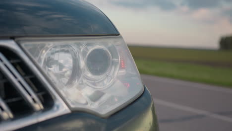close up of modern car headlight blinking beside quiet countryside road, shiny chrome details reflecting soft light from serene open field, under calm blue evening sky and distant trees