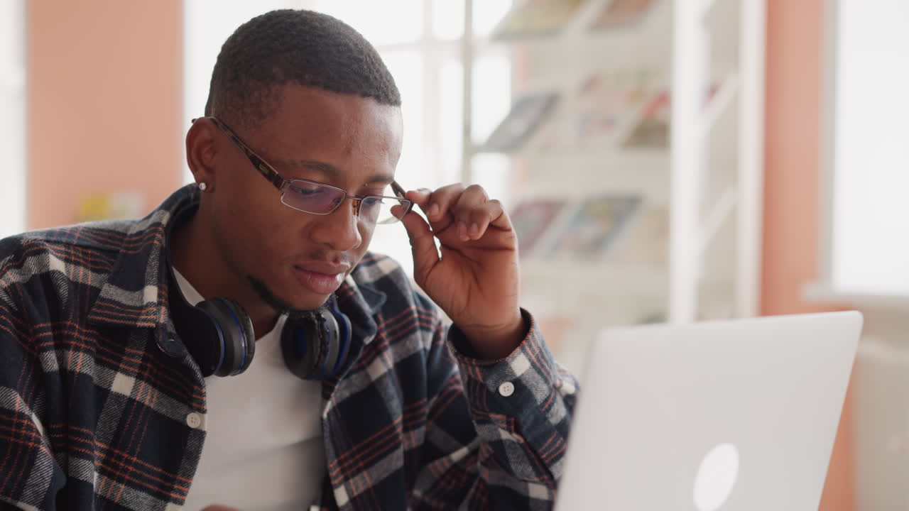 Student in glasses uses computer in library. African American man with earphones takes off glasses looking attentively at screen. Exams preparation at university