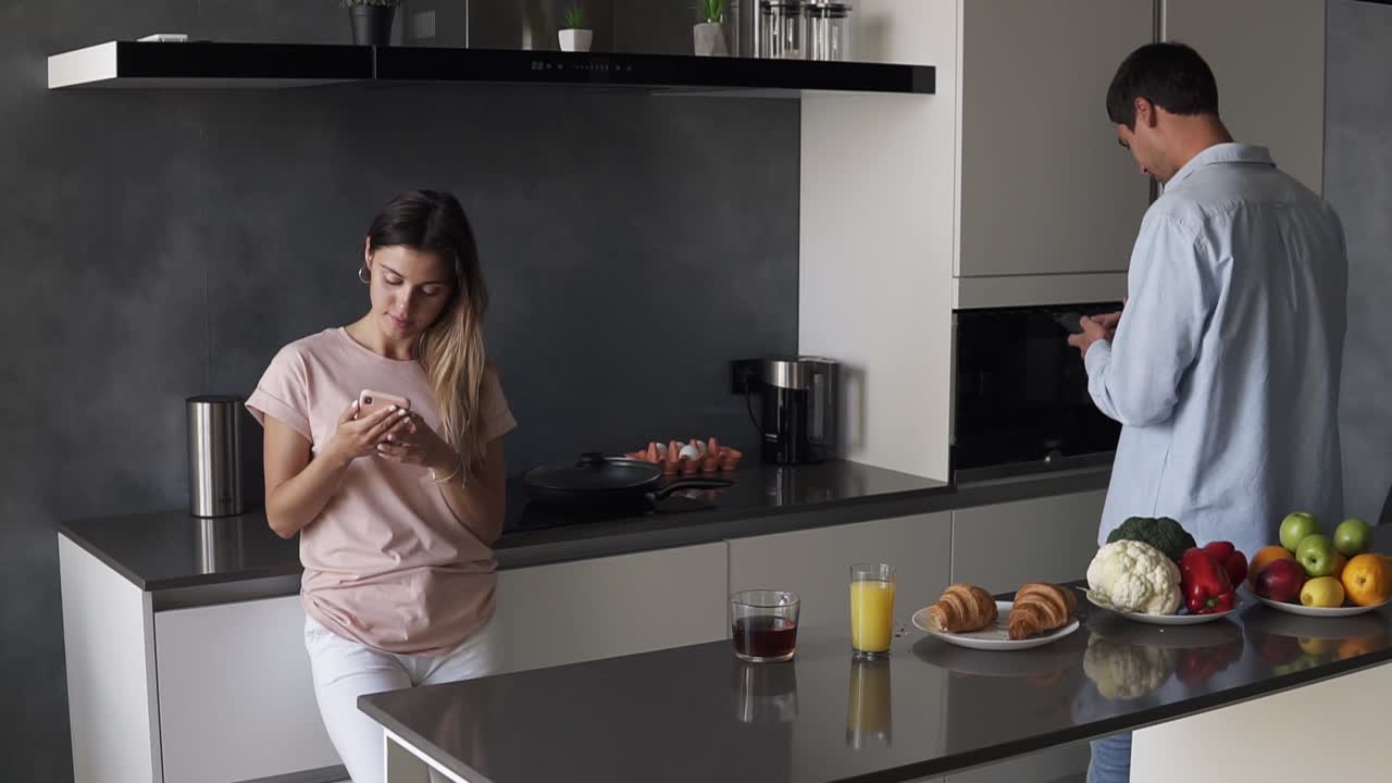 Young couple looking at something on their smartphones screen in the kitchen with modern loft design. Reading morning news or checking social media. Standing separately from each other