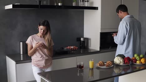 Pareja-Joven-Mirando-Algo-En-La-Pantalla-De-Su-Teléfono-Inteligente-En-La-Cocina-Con-Un-Diseño-Moderno-Tipo-Loft.-Leer-Las-Noticias-De-La-Mañana-O-Consultar-Las-Redes-Sociales.-Parados-Separados-Unos-De-Otros