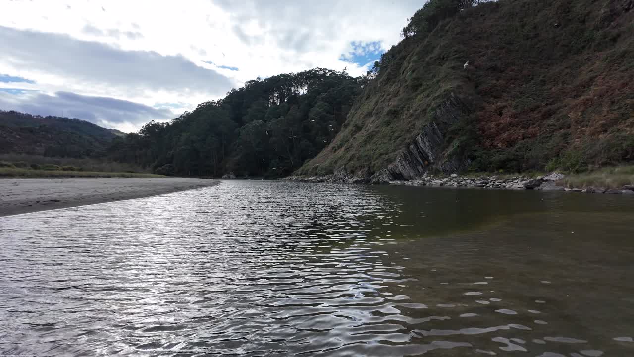 A low-angle view of rippling water meets a steep, rocky coastal cliff under an overcast sky
