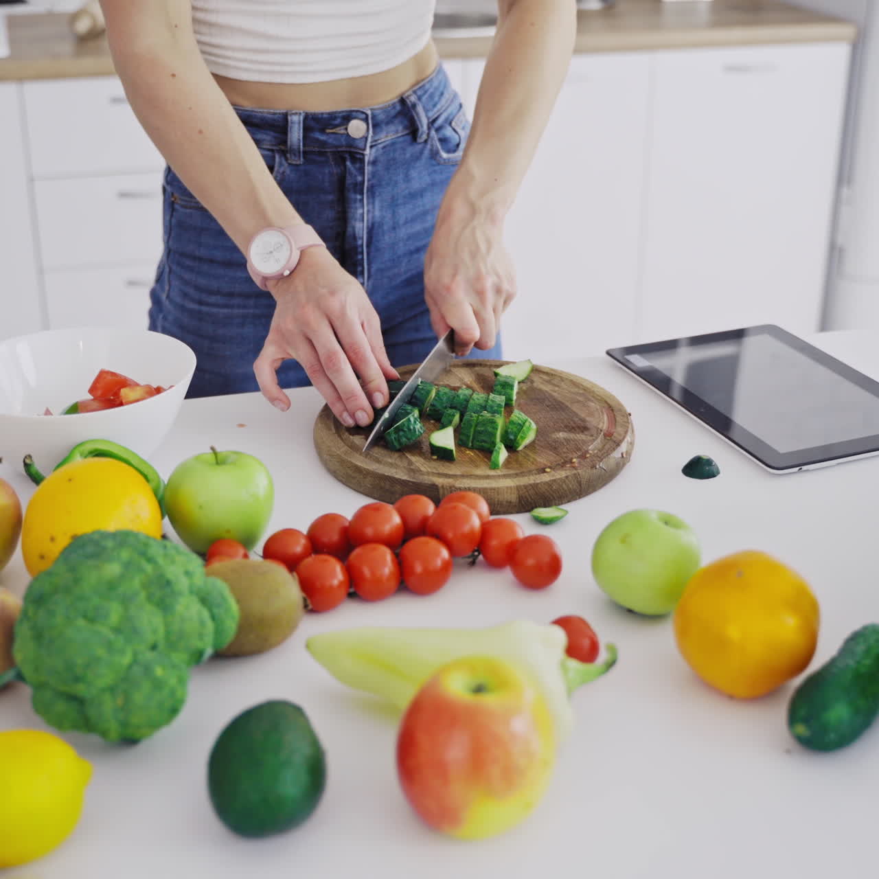 Delicious fruit and vegetables on a table and woman cooking. Housewife is cutting green cucumbers on a wooden board for making fresh salad in the kitchen.