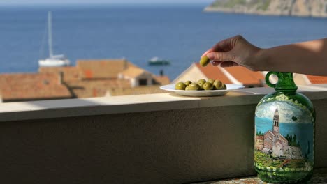 Woman-taking-olive-from-plate-on-terrace-with-sea-view,-Mediterranean-snack-dish