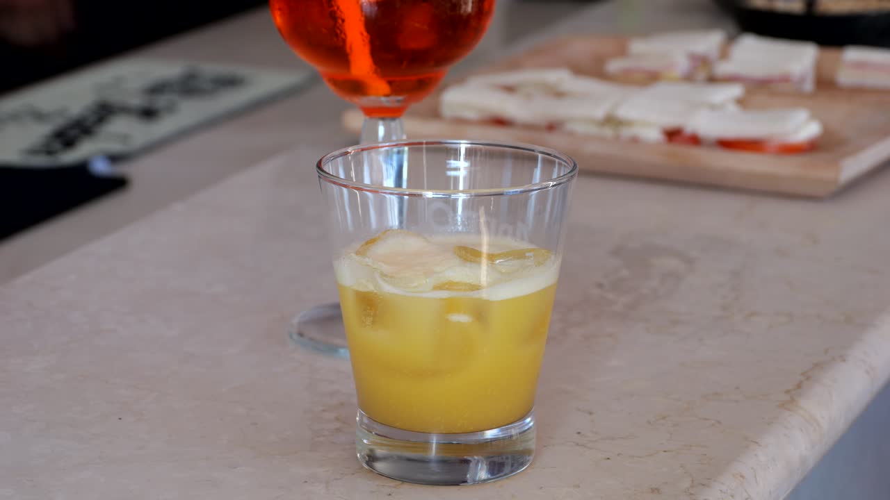 Glass of yellow cocktail served over ice on a bar counter with sliced appetizers in the background, captured by close-up shot