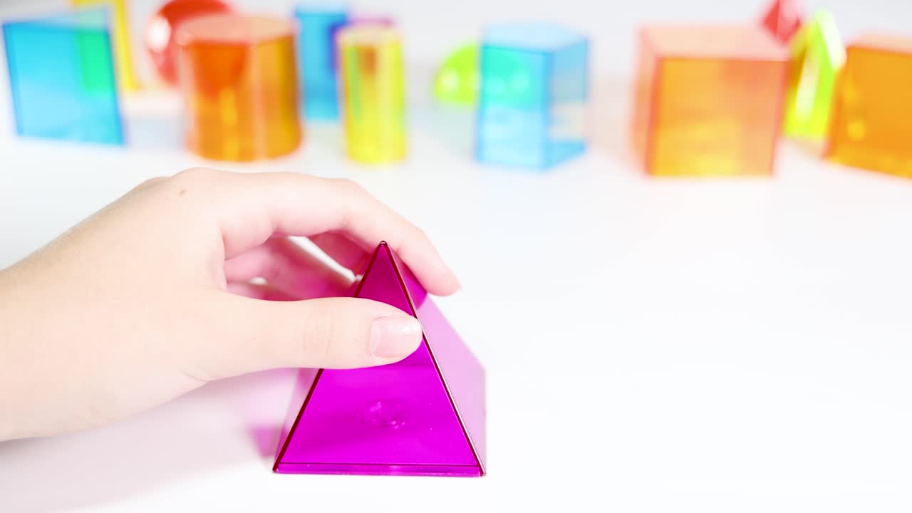 Hands manipulate vibrant 3D geometric shapes on a white surface, demonstrating educational concepts in a bright, playful setting