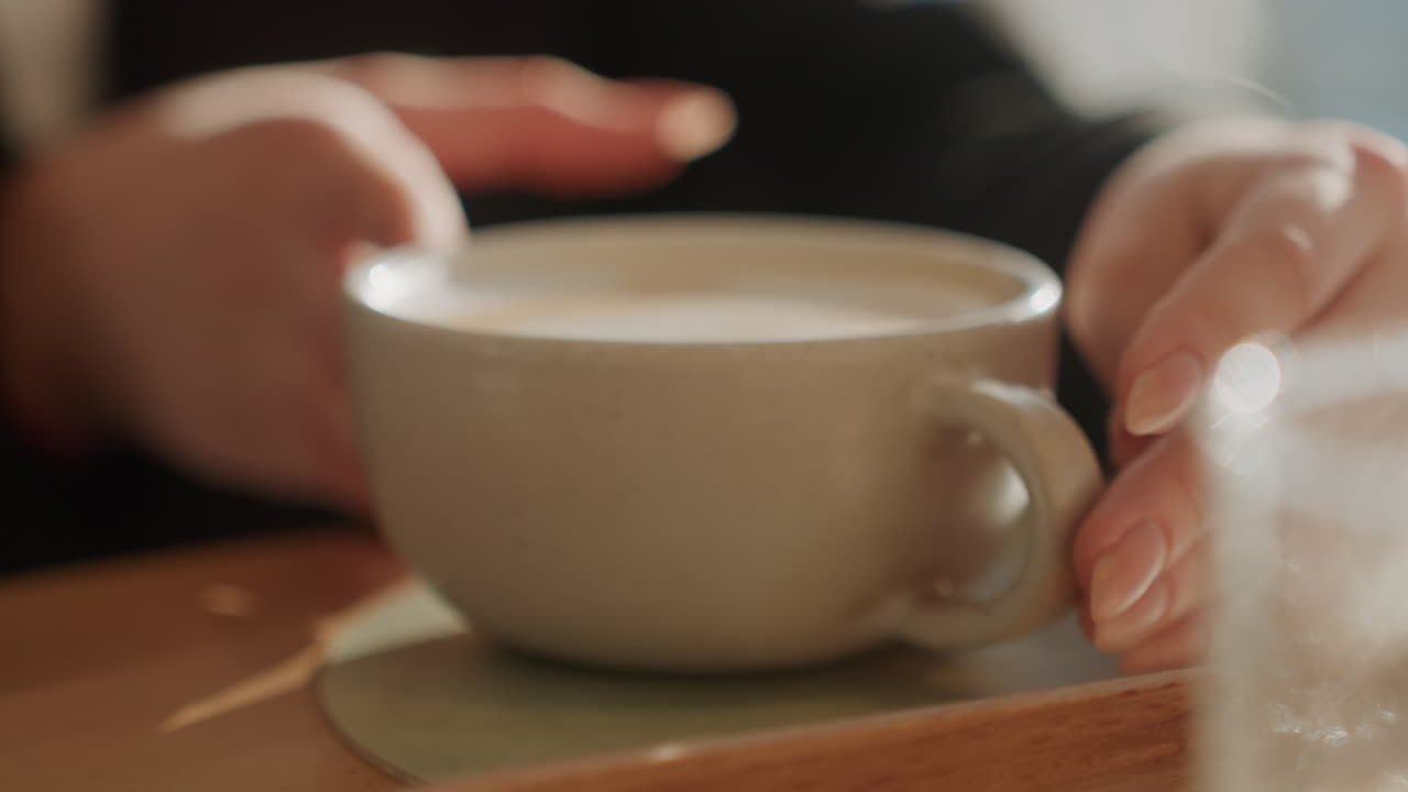 Mani calde in un caffè che stringono una tazza di latte, luce solare soffusa e il dettaglio della schiuma catturano il rituale calmo di un avventore che culla una tazza di ceramica su un tavolo di legno, gesti lenti e deliberati, crema vellutata, vapore sottile, luce naturale.