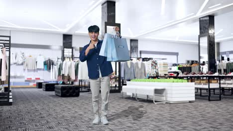 full view of an asian shopping man holding shopping bags up, pointing and smiling to camera while standing in clothing store