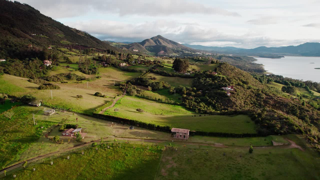 Filmato aereo con zoom in della campagna di Guatavita, che mostra campi verdi, case rurali e montagne distanti in una giornata di sole.