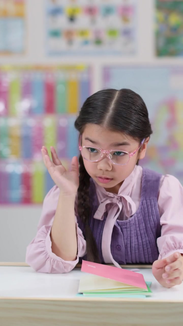 Una bambina con gli occhiali appare annoiata ed esausta mentre legge al suo banco in classe.