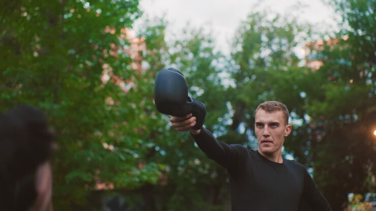 male athlete holding kettlebell outdoors with focused expression, park trees and dusk light framing slow controlled onearm hold, coachstyle demonstration of grip, core stability and functional