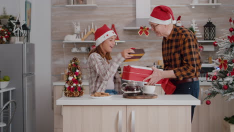 Fröhliche-Familie-Bringt-Weihnachtsgeschenkverpackung-Mit-Schleife-Darauf-In-Die-Weihnachtlich-Dekorierte-Küche