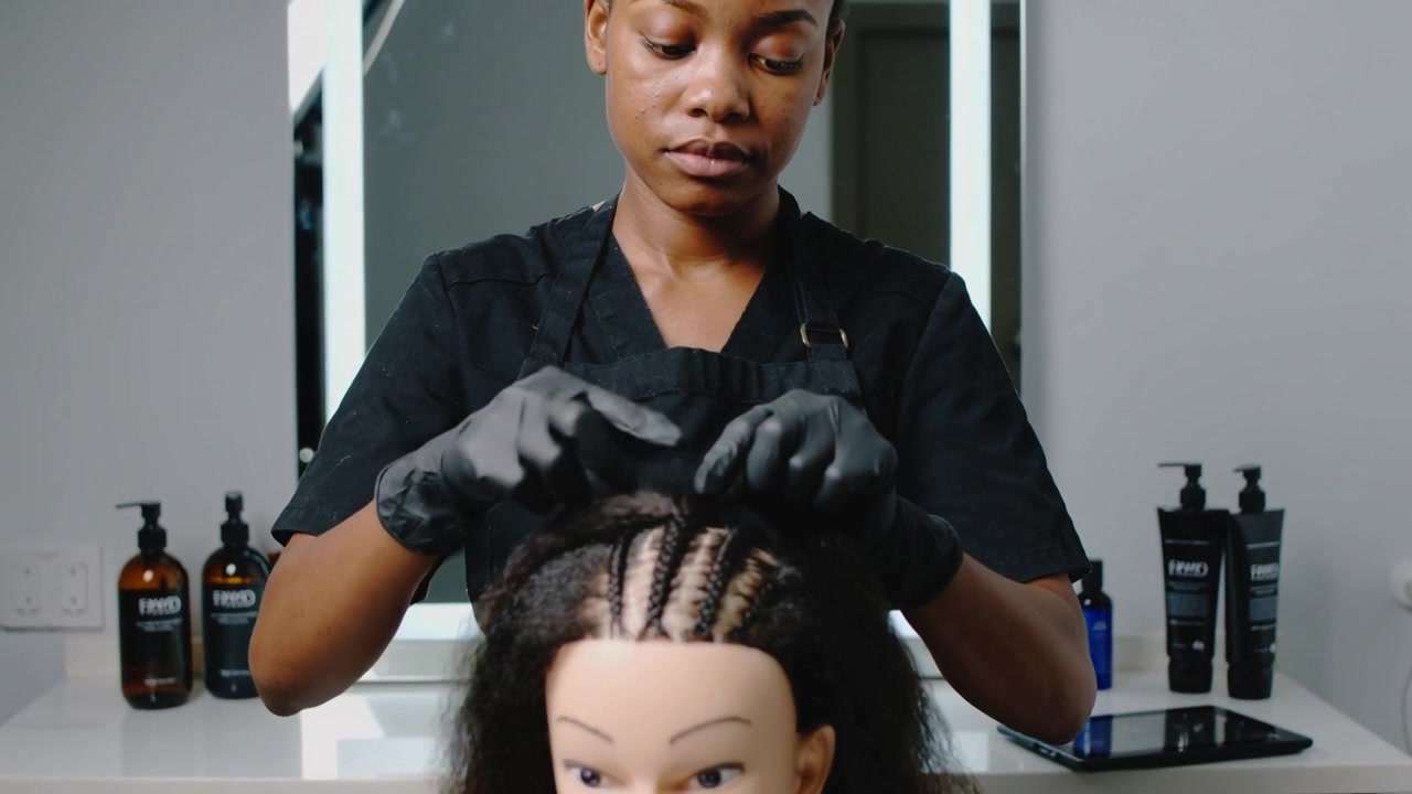 Braiding salon stylist pulling mid cornrow on mannequin after front rows set, training with gloves