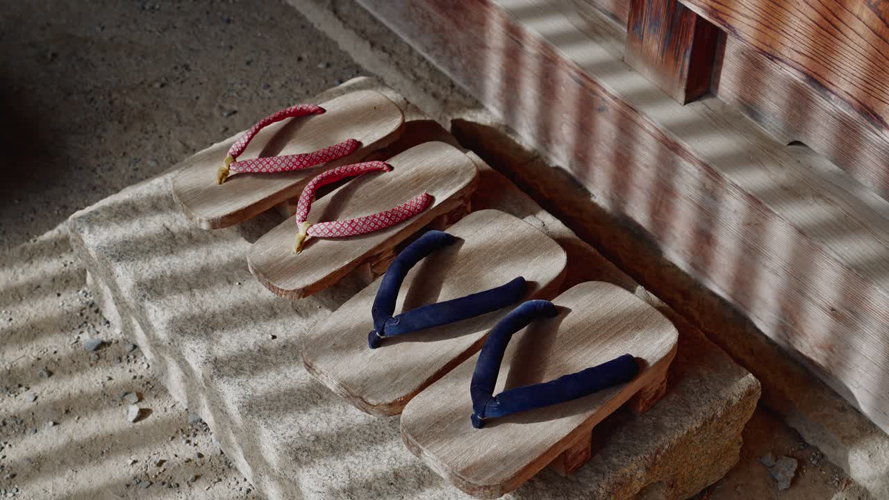 Twee paar houten geta-sandalen met rode en blauwe bandjes rusten op een stenen trede bij de ingang van een historisch huis. Ochtendzonlicht door een roosterdeur creëert een prachtig gestreefd schaduwpatroon.