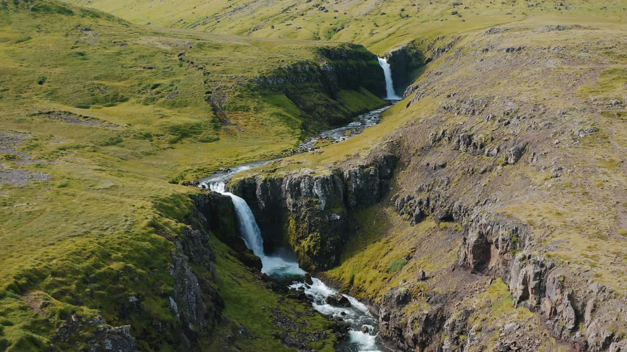 vuelo de avión no tripulado sobre una cascada que desciende en medio de las colinas verdes en islandia, en verano.