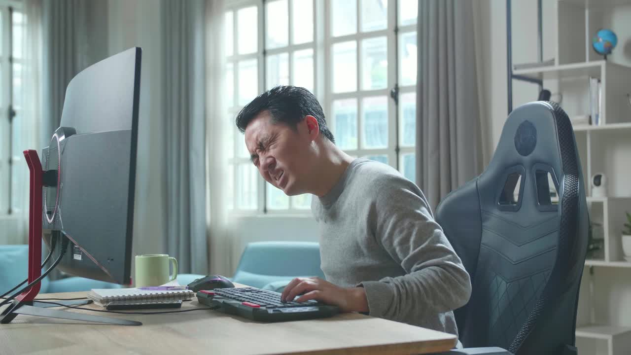 Asian Man In Long Sleeved T-Shirt Having Backache While Using Desktop Computer For Working At Home.