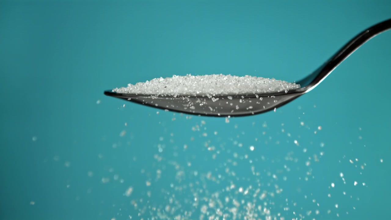 Zucchero semolato bianco che cade da un cucchiaio su sfondo verde acqua
